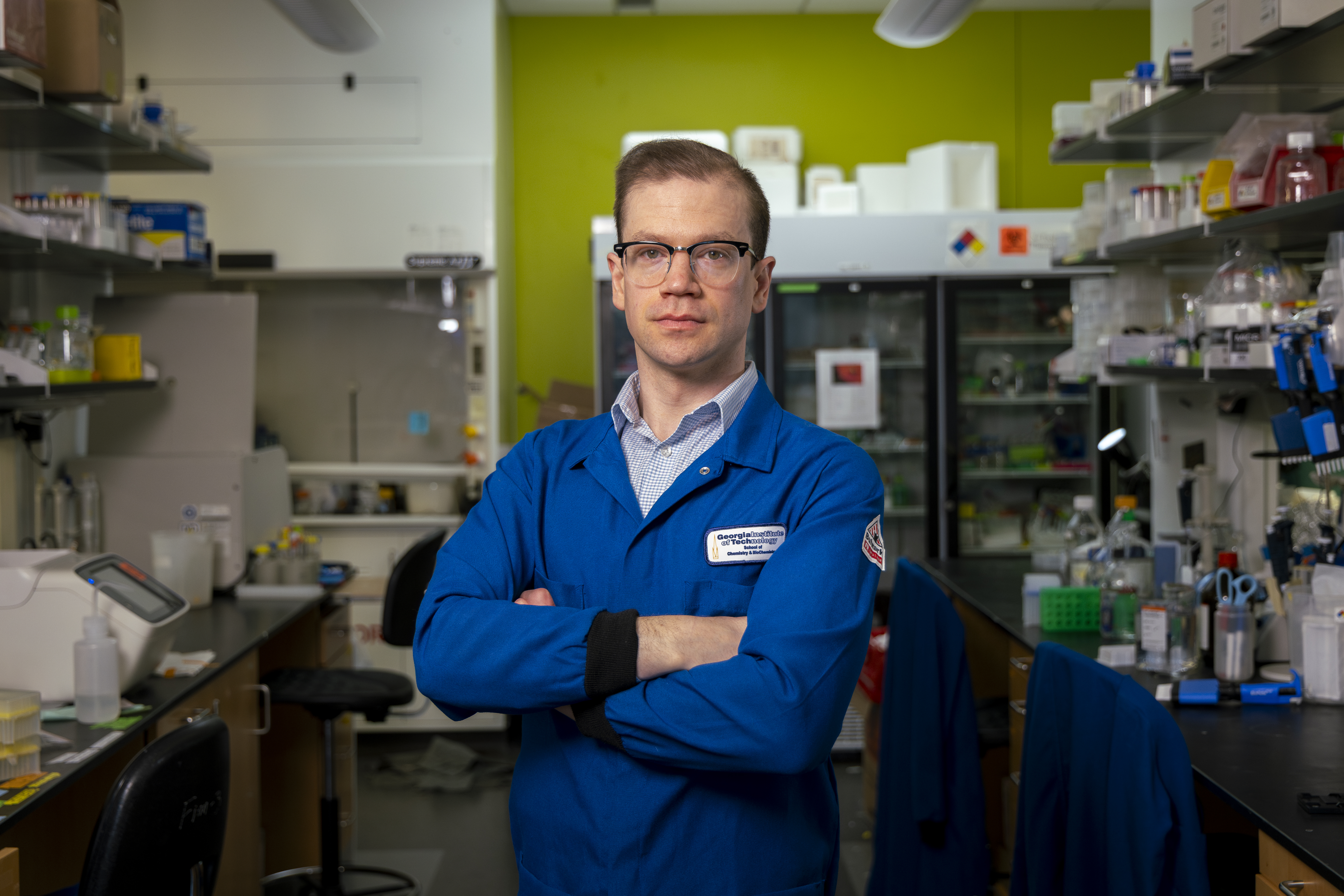 A person wearing a blue lab coat stands with arms crossed in a laboratory filled with shelves of scientific equipment, supplies, and a refrigerator unit in the background.
