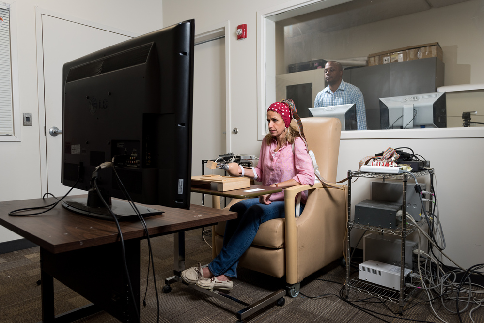 <p>Lewis Wheaton (back) directs Georgia Tech’s Cognitive Motor Control Lab.</p> A person seated in a beige chair using a computer setup with multiple cables and devices, facing a large monitor in a testing or research room, with another individual visible through a window in an adjacent control room.